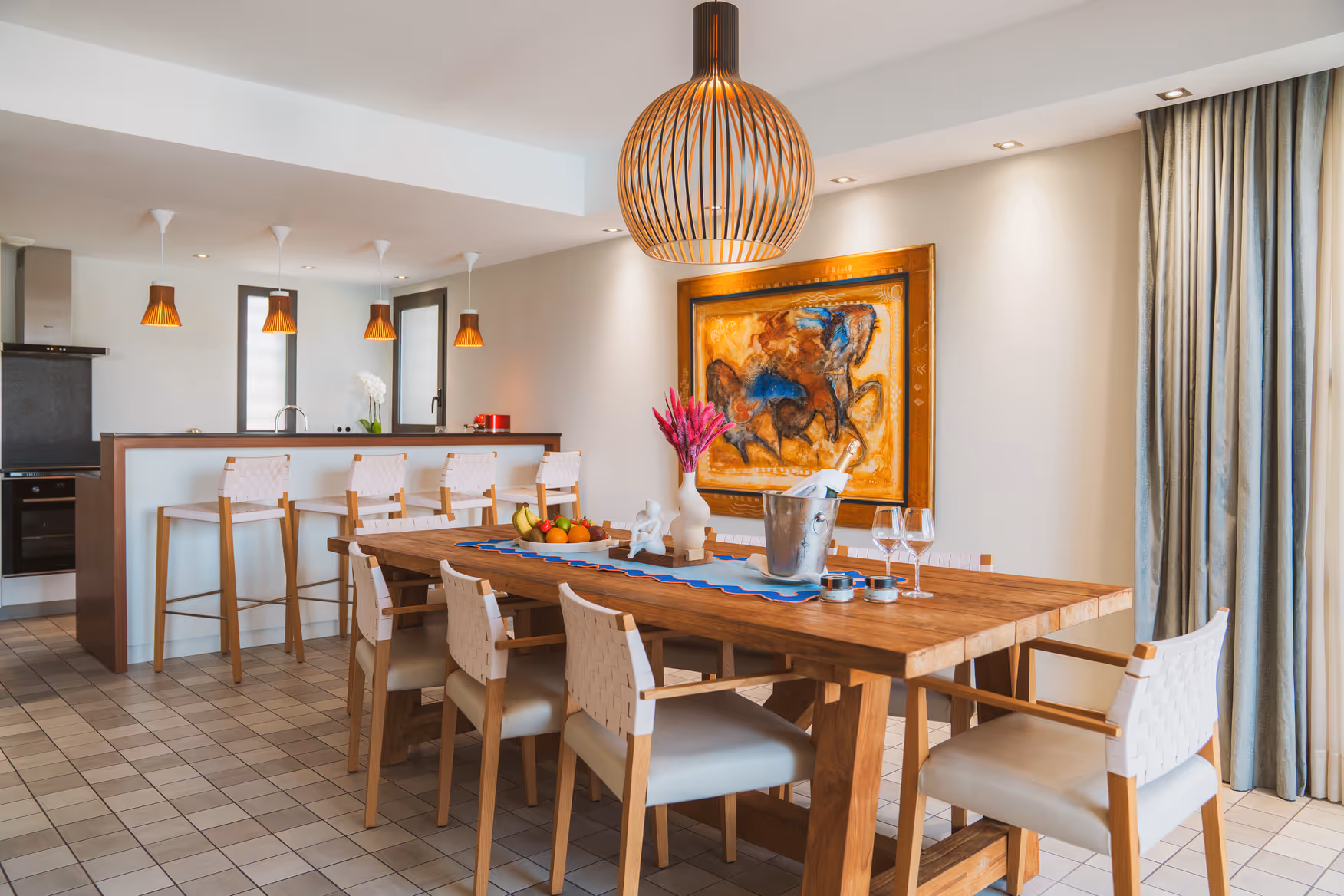 Salle à manger moderne avec grande table en bois, huit chaises blanches, suspensions en bois, et une peinture colorée accrochée au mur.